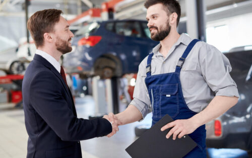 Side view portrait of smiling handsome businessman shaking hands with bearded mechanic standing in car service and repair center, copy space