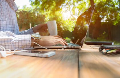 Smart casual dressed person working on computer drinking coffee mug sitting at rough natural wooden desk outdoor with green tree and sun on background